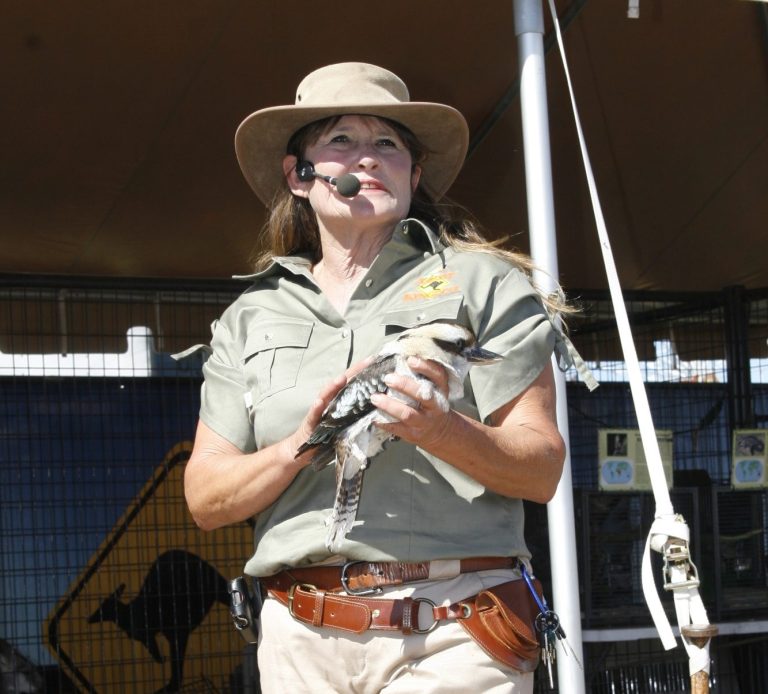 Woman in safari outfit holding a bird, speaking into a headset microphone outdoors.